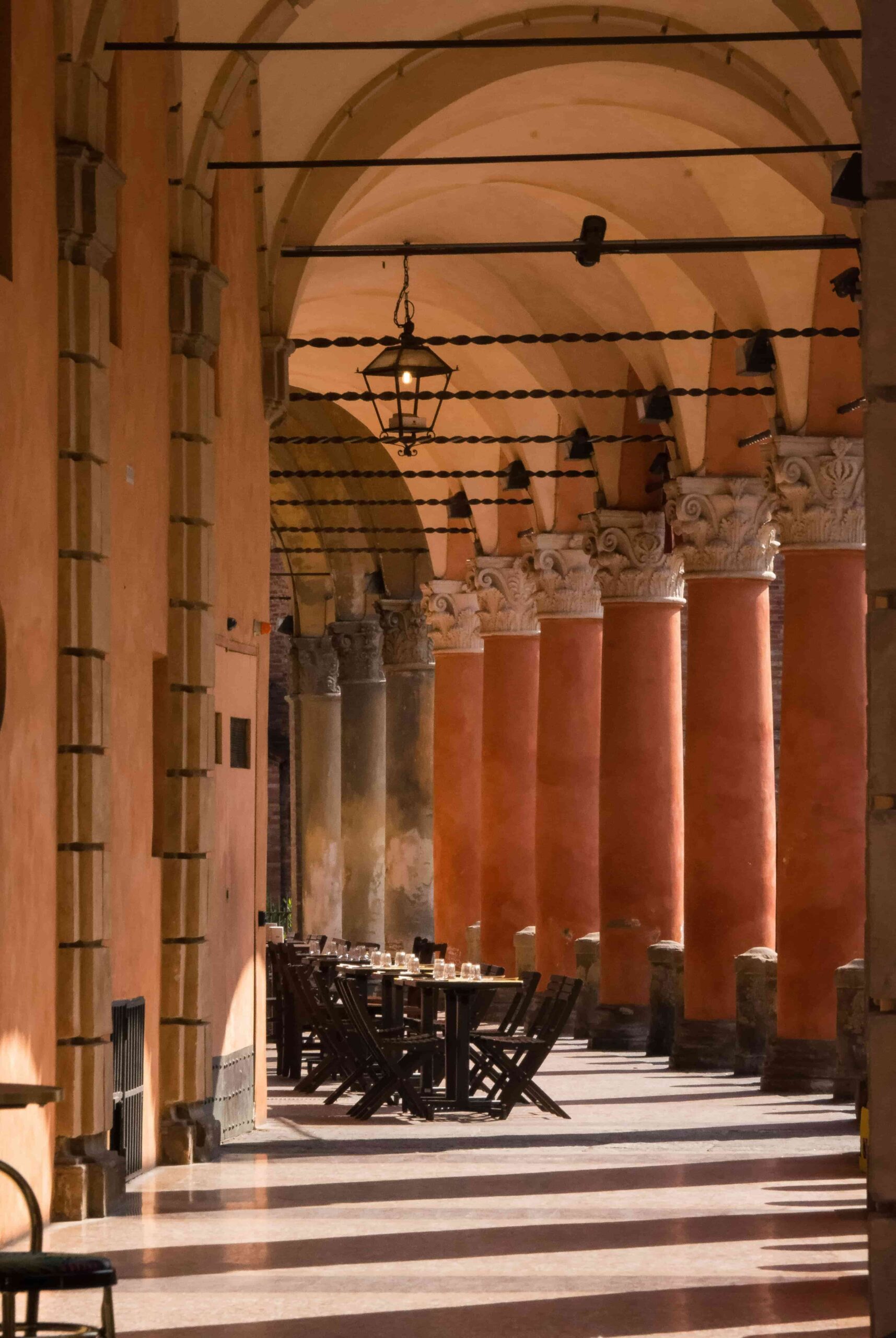 A long, covered walkway under Bologna’s iconic porticoes, with golden sunlight casting shadows on the cobblestone path. A few pedestrians walk beneath the historic archways, showcasing the city’s UNESCO-listed architecture.