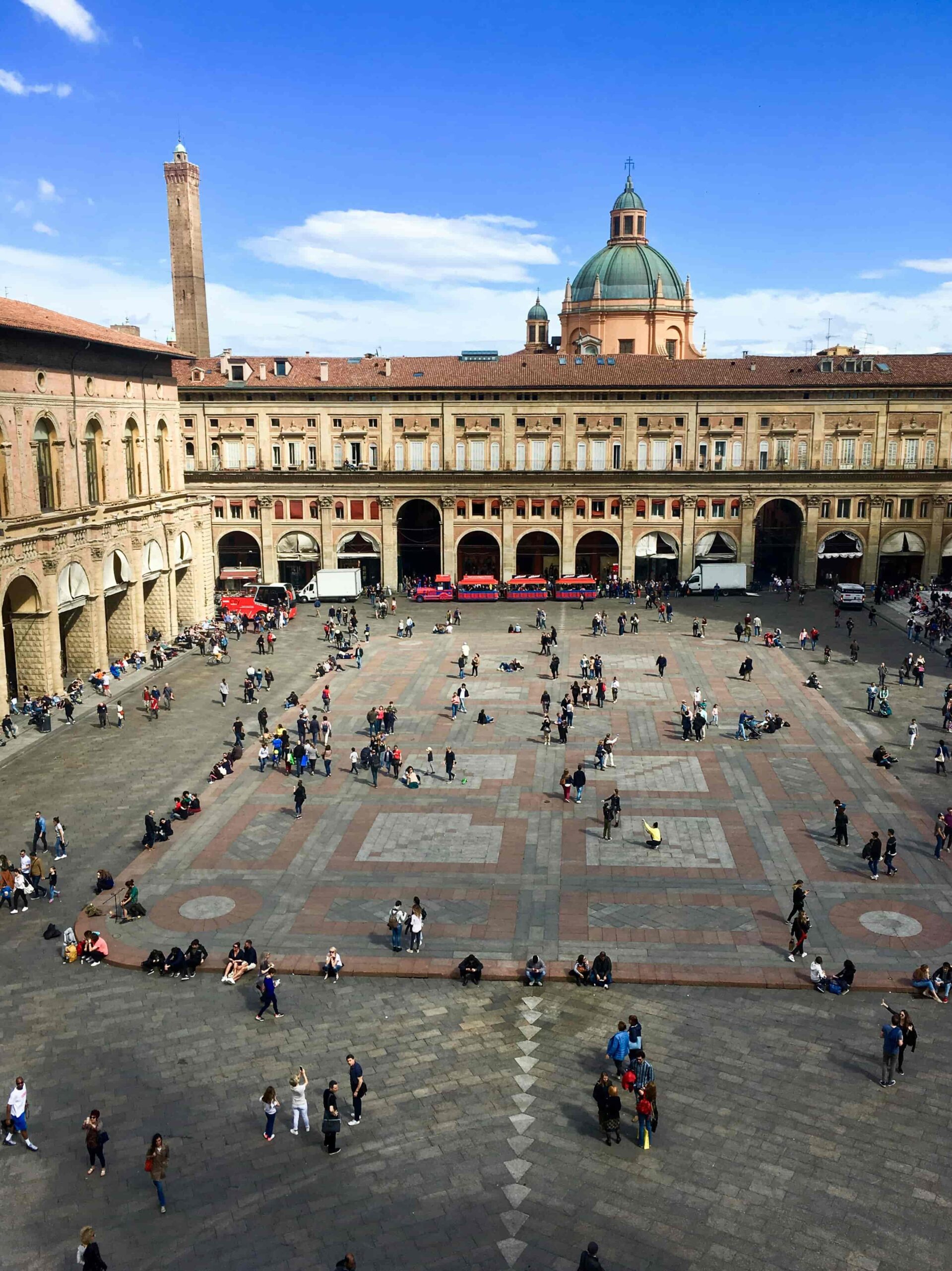 Breedhoekzicht van Piazza Maggiore in Bologna, Italië, bij zonsopgang. De basiliek van San Petronio staat op de achtergrond, badend in warm ochtendlicht, met een paar mensen die door het plein wandelen.