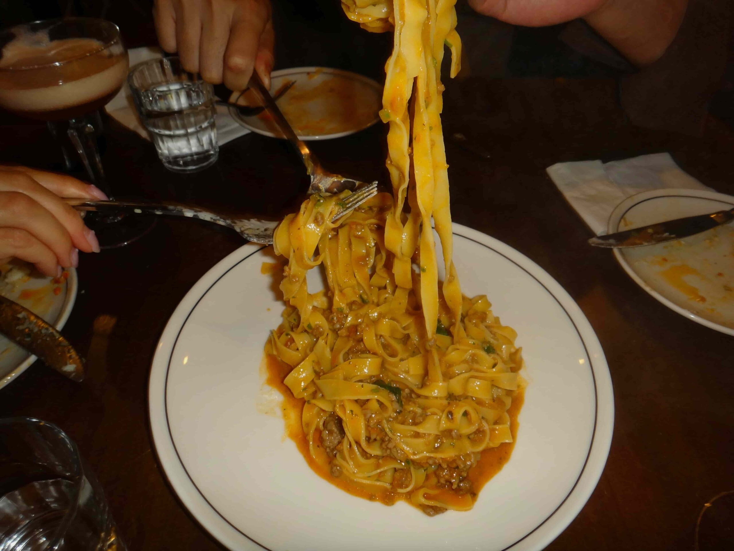 En närbild av en tallrik Tagliatelle al Ragù, toppad med parmesanost, som sitter på ett rustikt träbord. Ett glas rött vin, färskt bröd och oliver följer med rätten i en mysig italiensk trattoria.