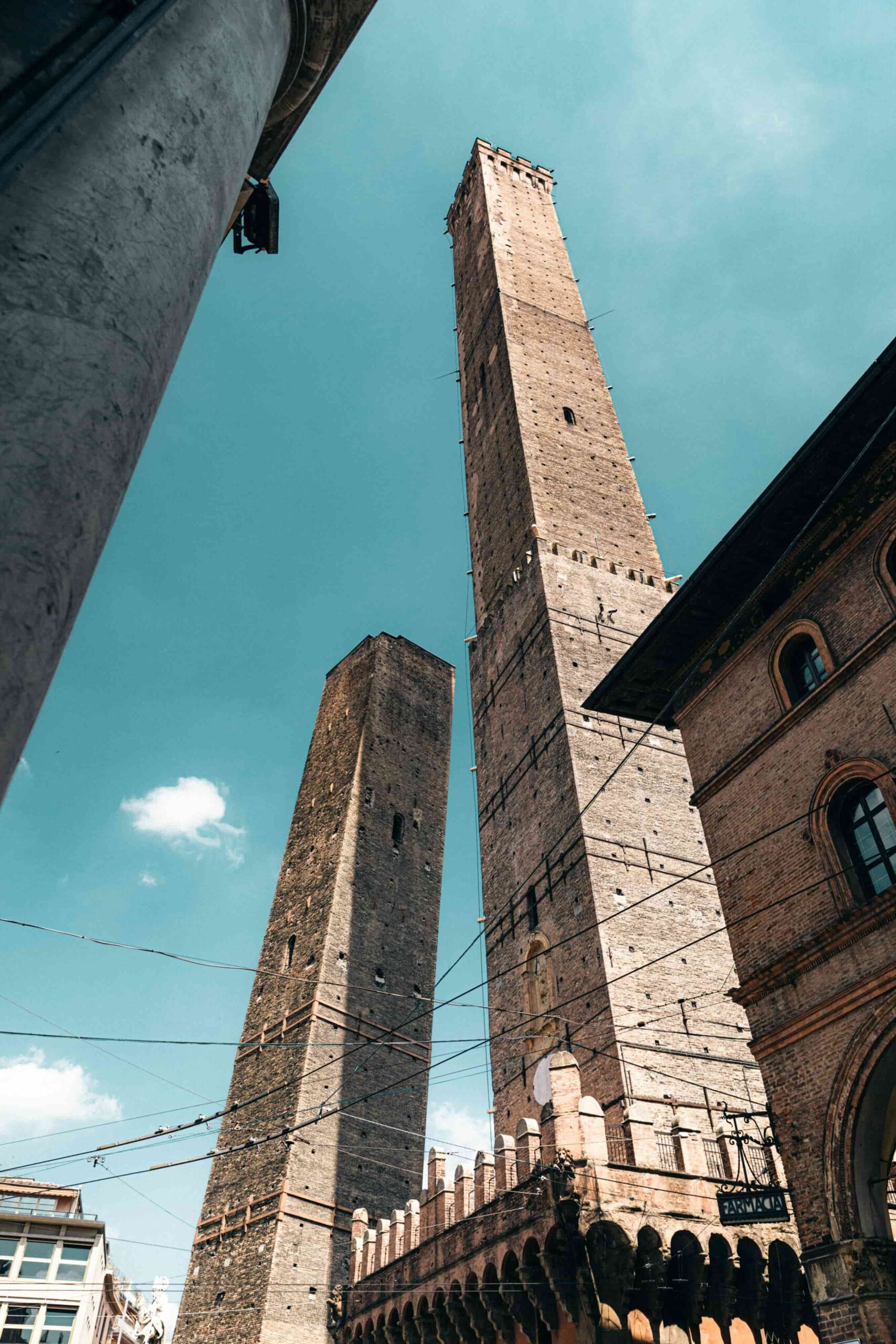 Ein dramatischer Aufwärtsschwung der Zwei Türme von Bologna, Asinelli und Garisenda, die gegen einen klaren blauen Himmel stehen. Die historischen Backsteinstrukturen lehnen sich leicht, während Fußgänger darunter die mittelalterliche Architektur bewundern.