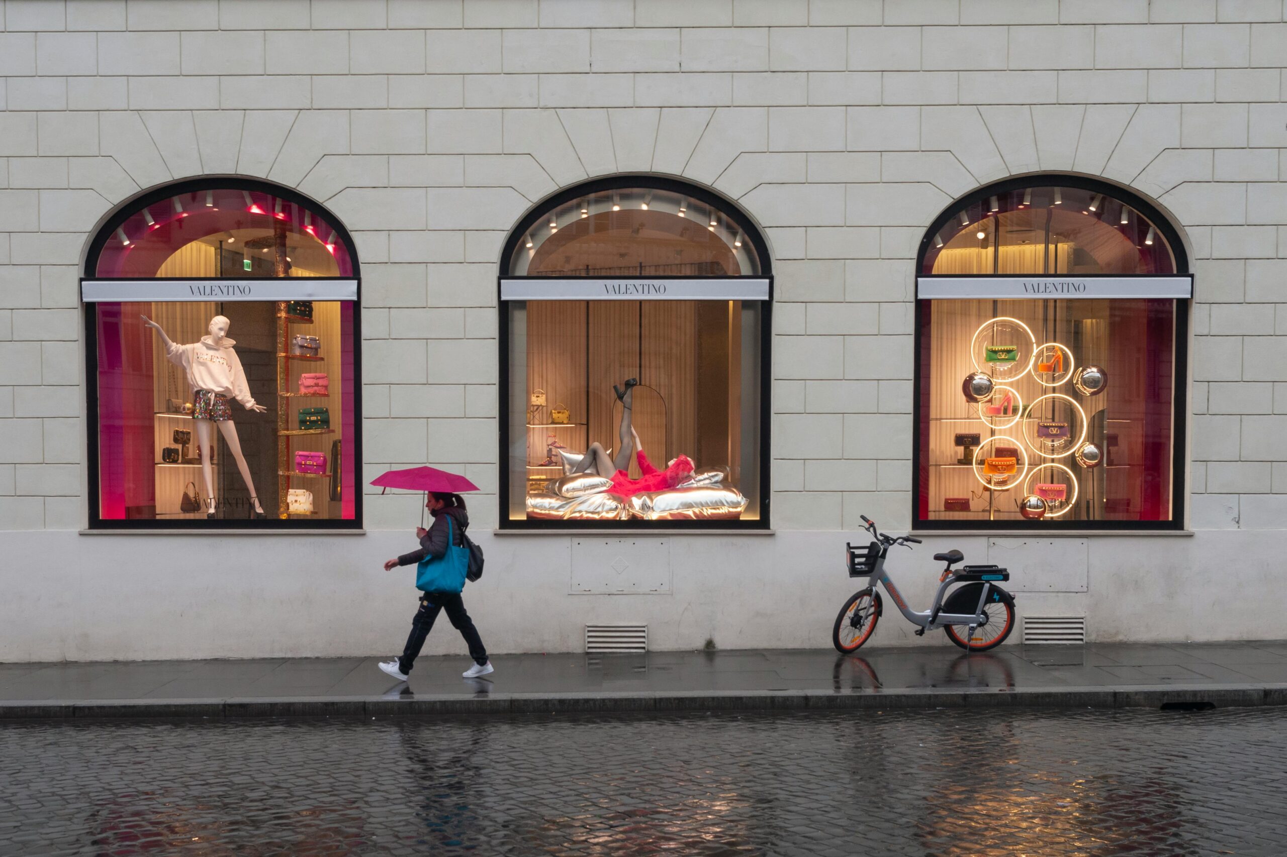 Person with a red umbrella walking along a wet street past shop windows