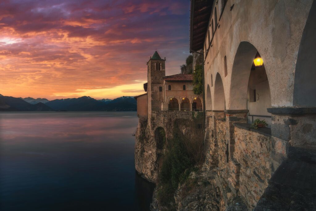 Scenic view of a historic monastery perched on a cliff overlooking Lake Maggiore at sunset in Italy.